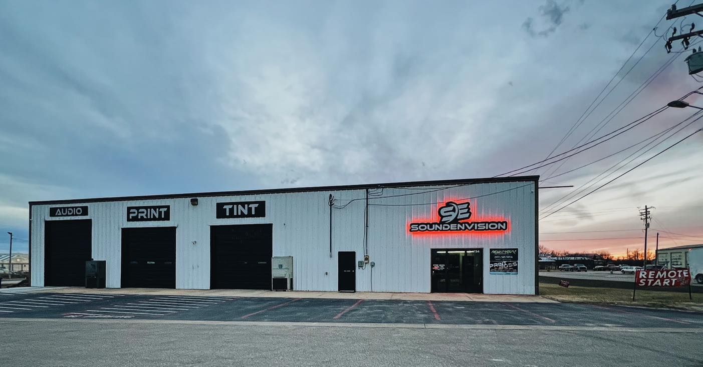Small white auto shop with open bays and red neon sign at dusk under a cloudy sky