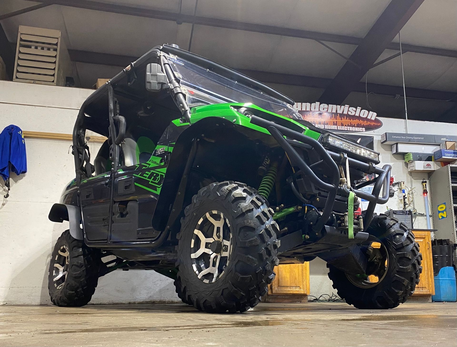 Green and black off-road side-by-side vehicle in a garage, with large knobby tires and lifted suspension