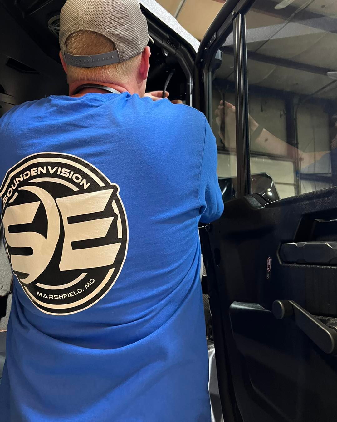 Person in blue shirt with a circular logo stands beside an open vehicle door in a garage.