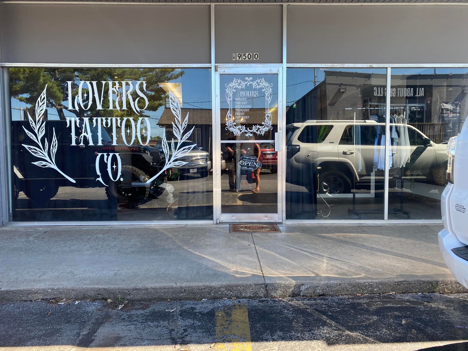 Storefront with “Lovers Tattoo Co” sign and glass door on a street, reflected white SUV outside