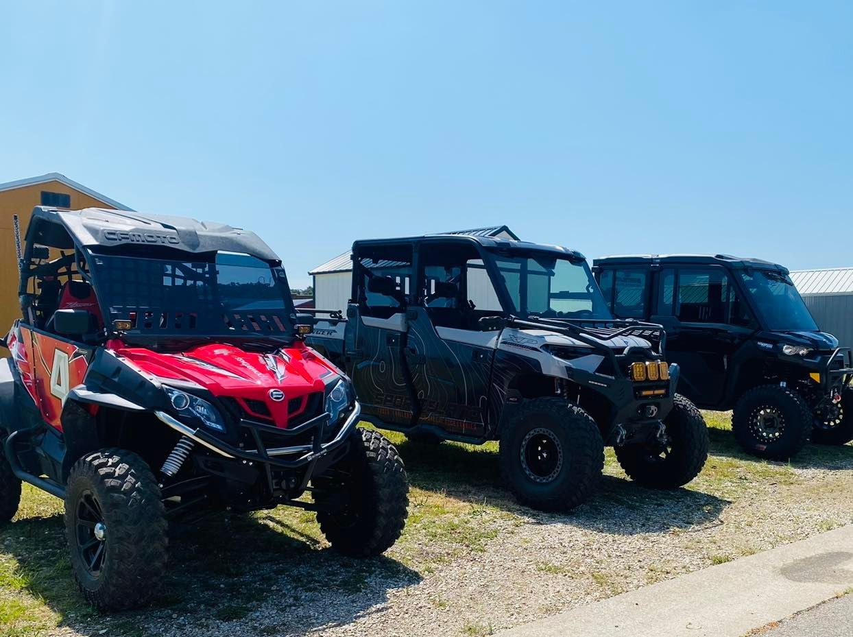 Three off-road UTVs parked on gravel under a clear blue sky, including a red one in front.