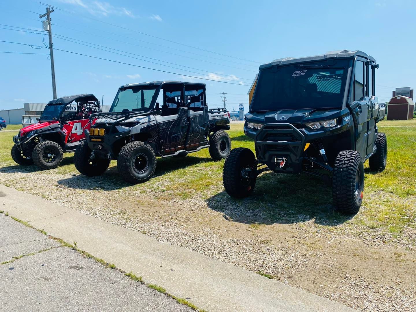 Three side-by-side off-road utility vehicles parked on gravel beside a road under a blue sky.
