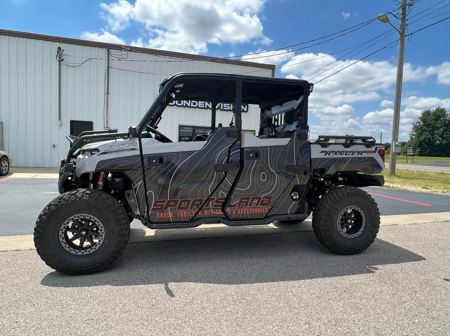 Black lifted off-road side-by-side parked on pavement near a white building under a blue sky