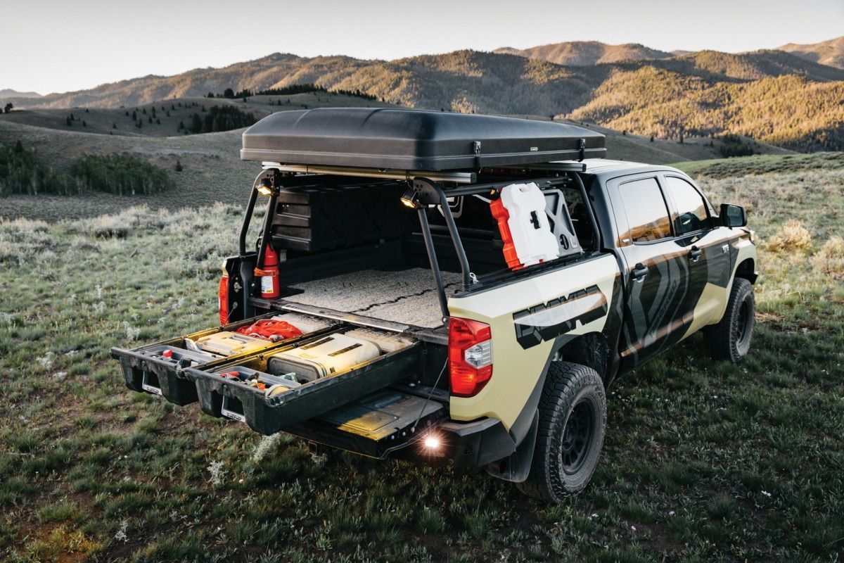 Black pickup with camper setup parked on grassy field at sunset, tailgate open with gear and roof storage.