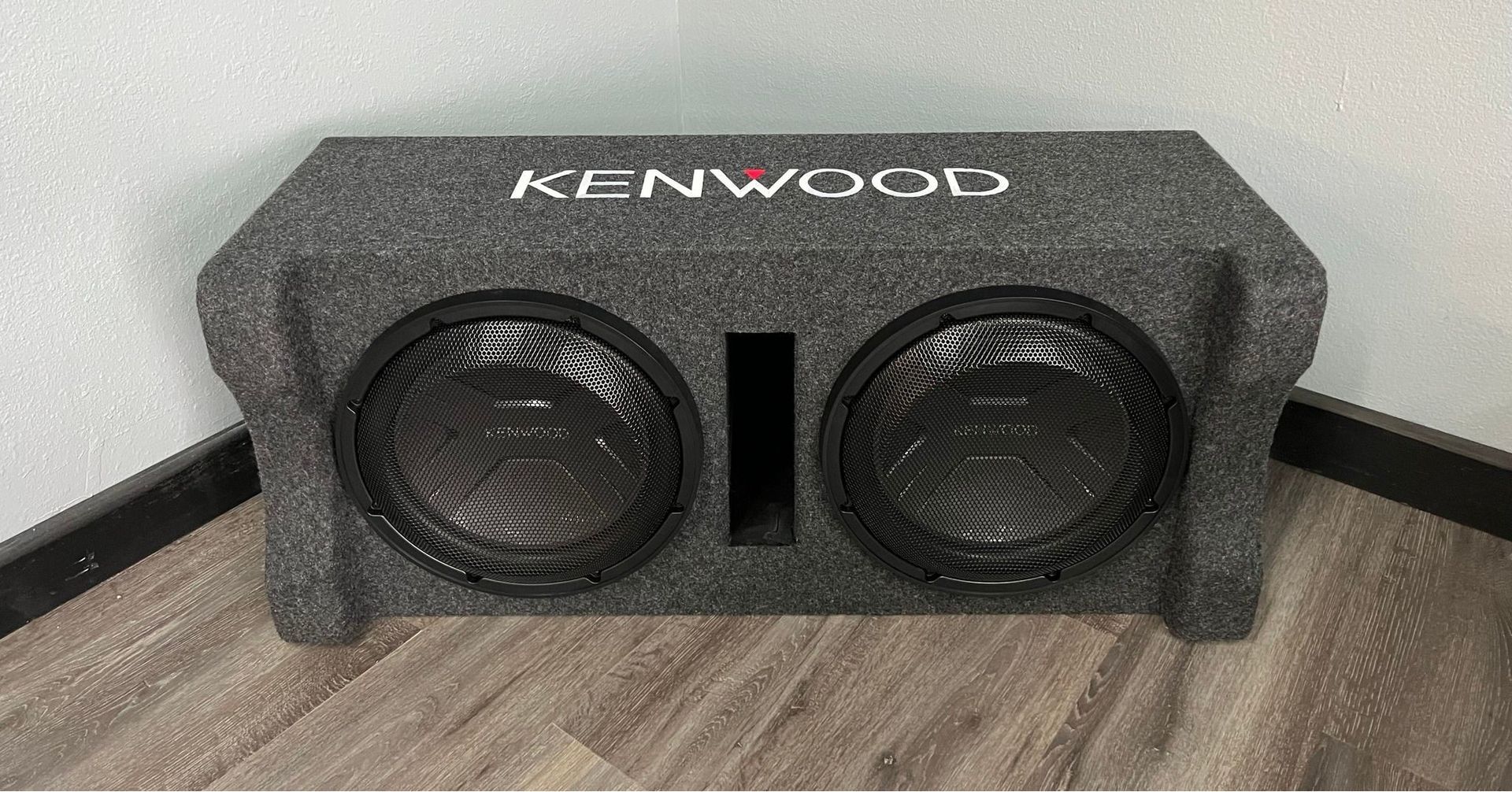 Two Kenwood subwoofers in a gray carpeted box on a wooden floor, with a black wall behind.