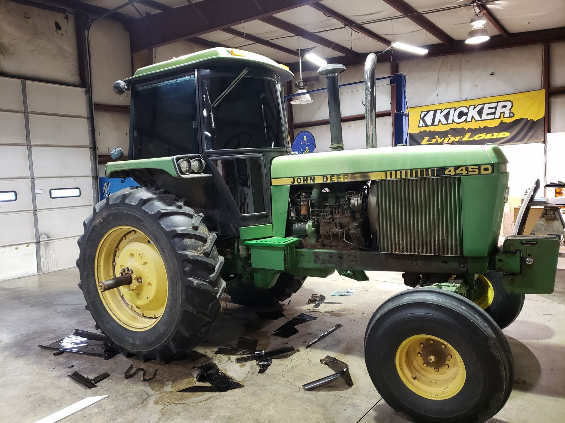 Green tractor with exposed engine in a garage, surrounded by scattered black parts and tools.