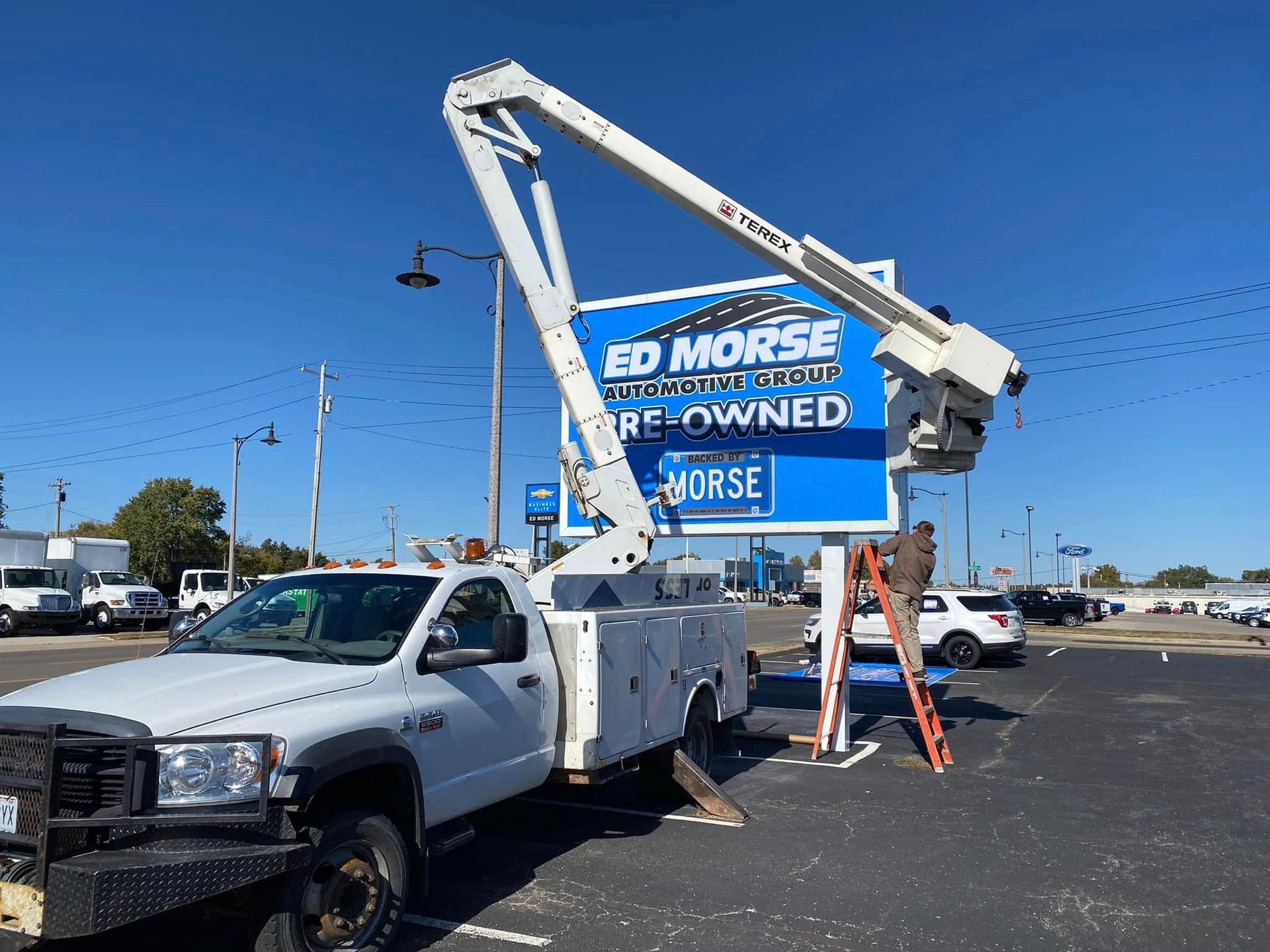 White utility truck with raised boom lift beside a used car lot sign under a blue sky.