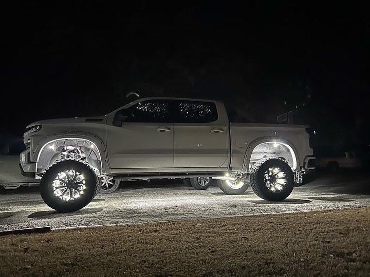 Silver lifted pickup truck parked at night on a gravel lot under bright lights