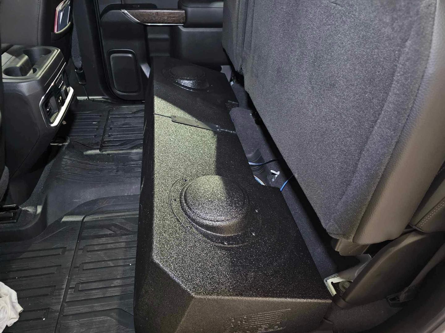 Black speaker box with round grille mounted on a vehicle floor beside a gray seat.