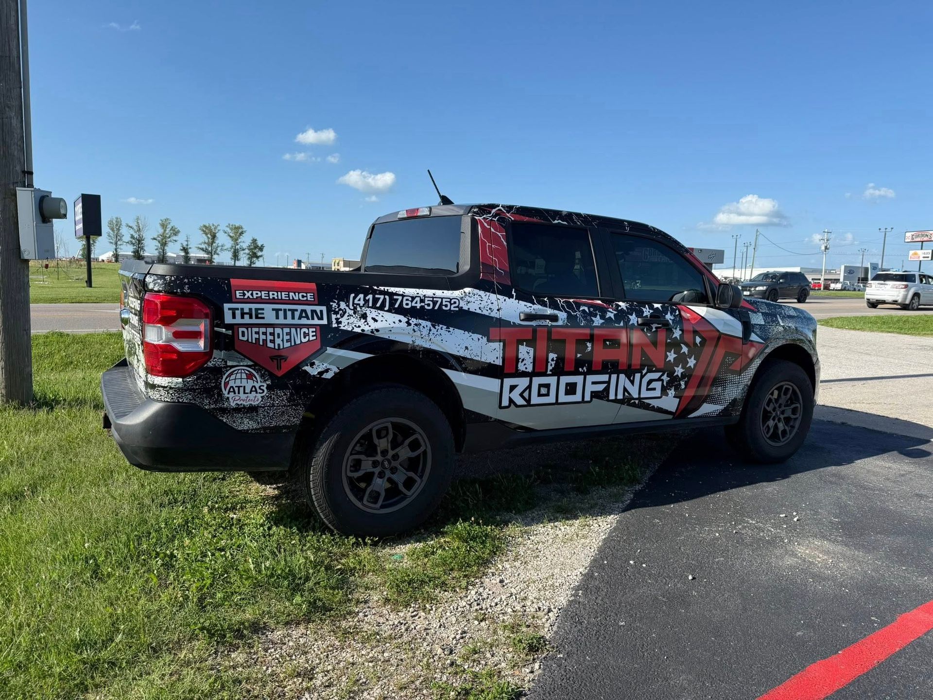 Red and black Titan Roofing pickup truck parked on grass by a gravel lot under a blue sky.