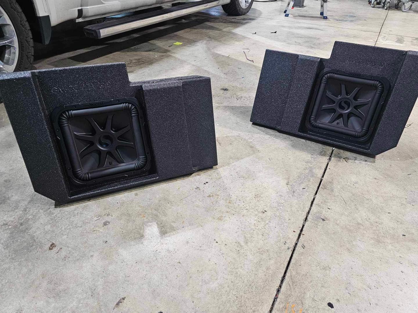 Two black subwoofers or speakers lying on a concrete garage floor beside a parked vehicle.