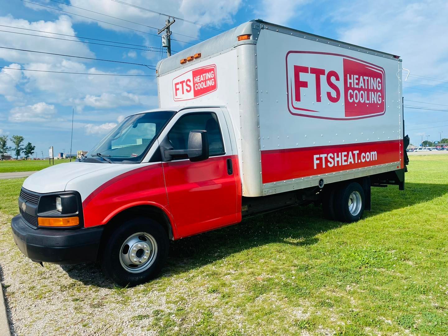 Red and white FTS Heating & Cooling box truck parked on grass under a blue sky.