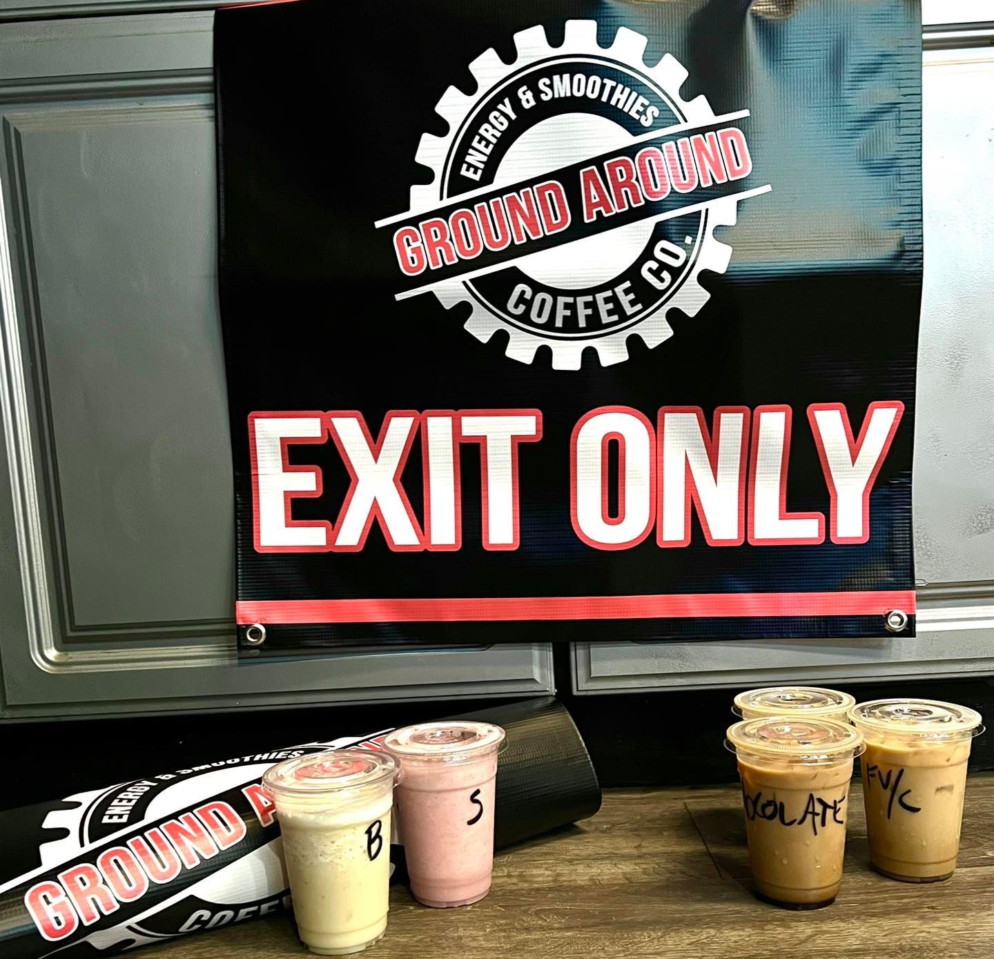 Coffee shop sign reading “EXIT ONLY” above four iced drinks on a wooden counter.