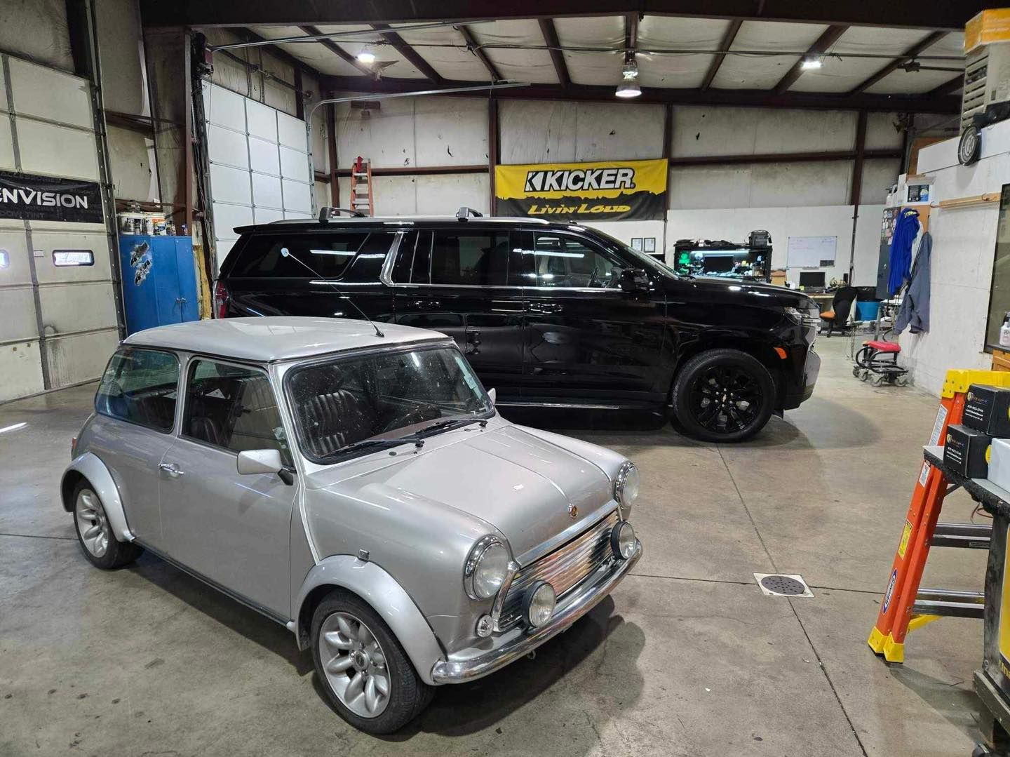 Two cars in an auto shop, including a silver classic Mini in the foreground and a black SUV behind it.