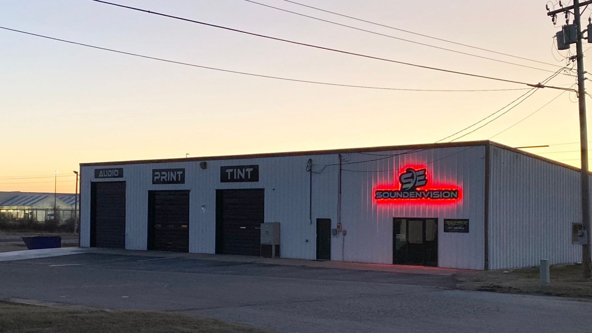 Small industrial building at dusk with three garage doors and a red neon sign glowing on the front.