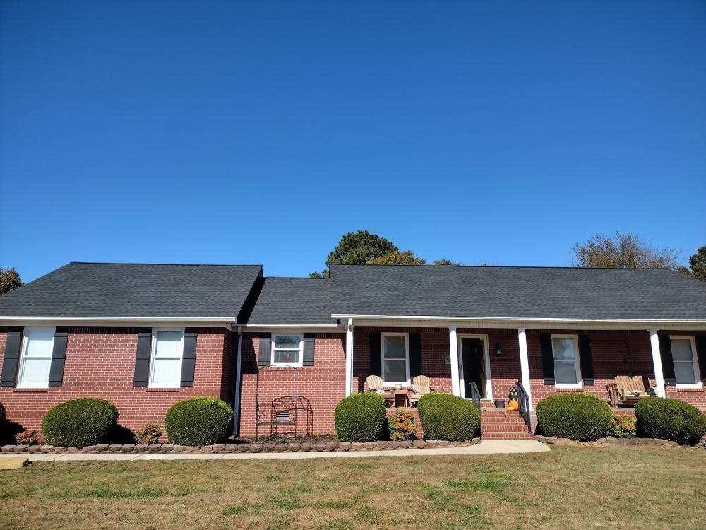 Brick house with a dark roof and white trim under a clear blue sky. Green shrubs in front.