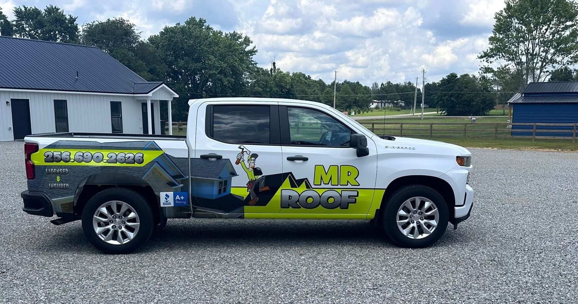 White MR Roof company pickup truck parked on gravel driveway. Buildings and trees in background.