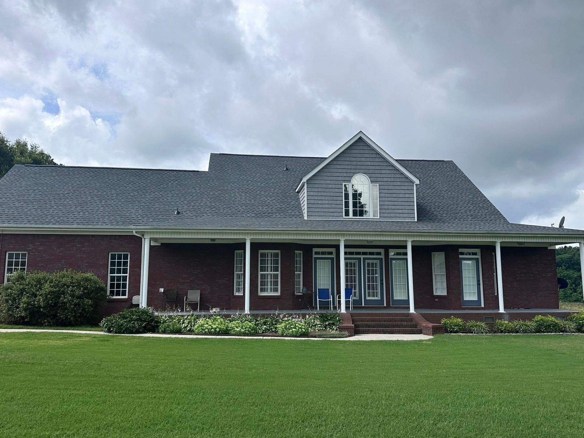 Brick house with gray roof, porch, and a dormer. Lawn in foreground, cloudy sky above.