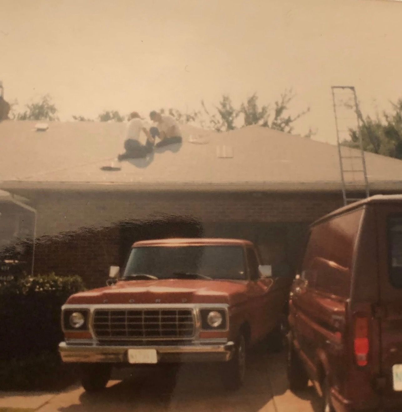 Two people on a roof working, with a red pickup truck and a van parked in front of a house.