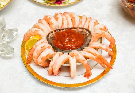 A shrimp cocktail platter featuring arranged cooked shrimp, a center bowl of red sauce, and a lemon wedge on a gold plate.