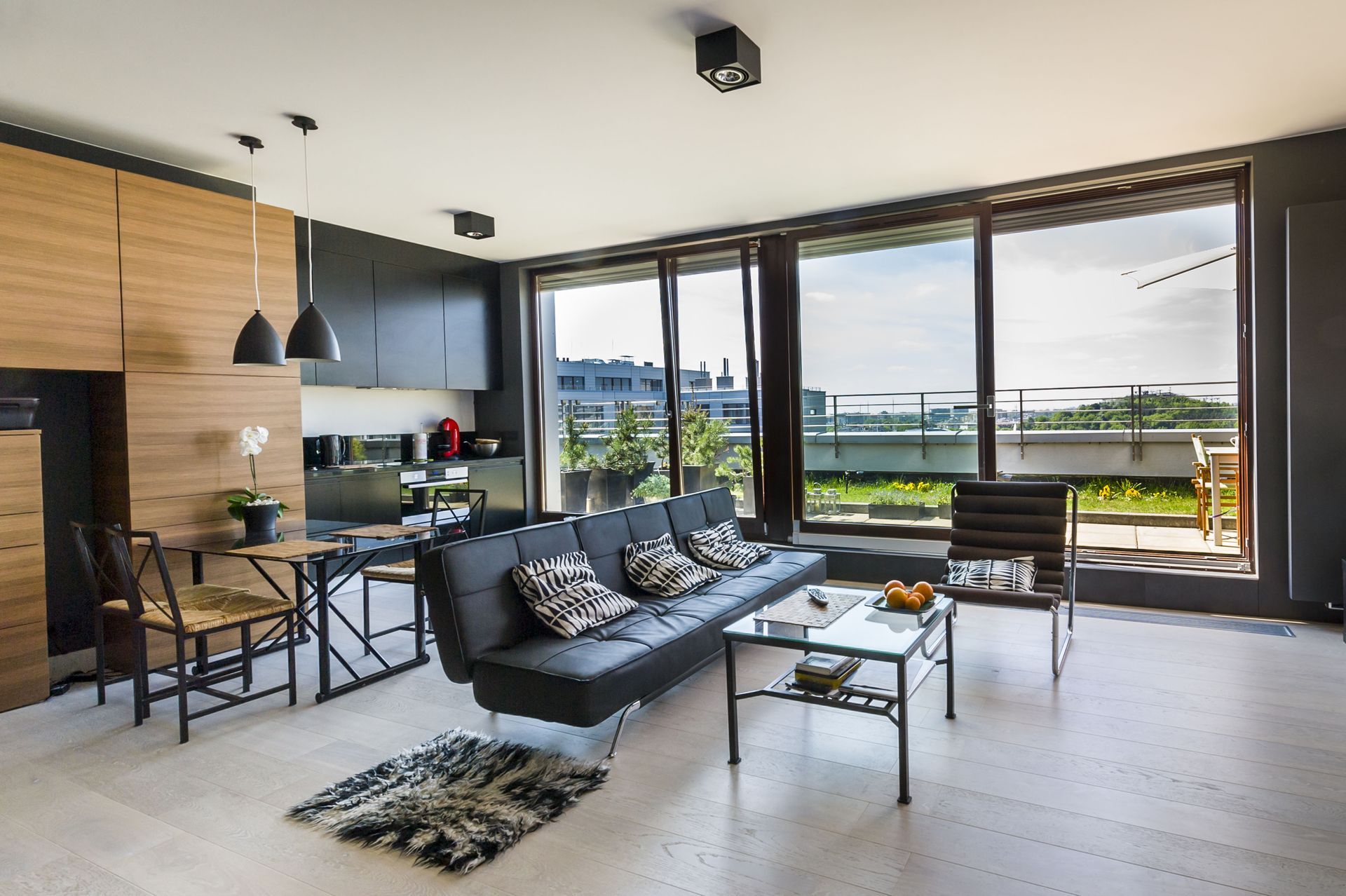 Modern living room with black sofa, kitchen area, and large windows overlooking a waterfront.