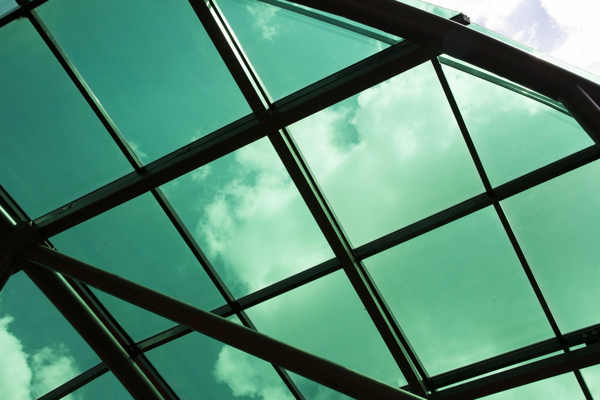 Green-tinted glass roof with black metal framework and cloudy sky beyond