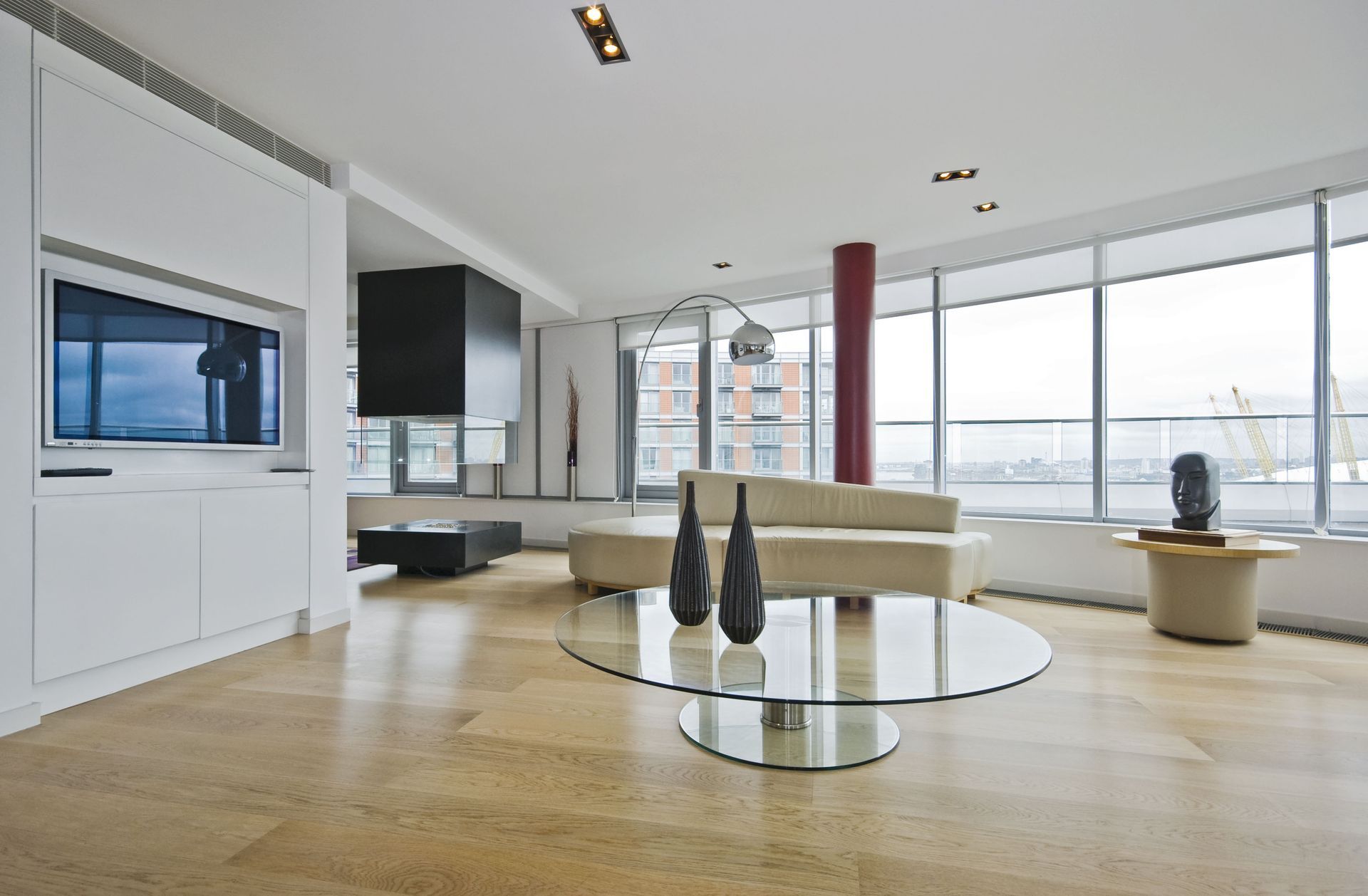 Bright modern living room with large windows, beige sofa, glass coffee table, and wall-mounted TV