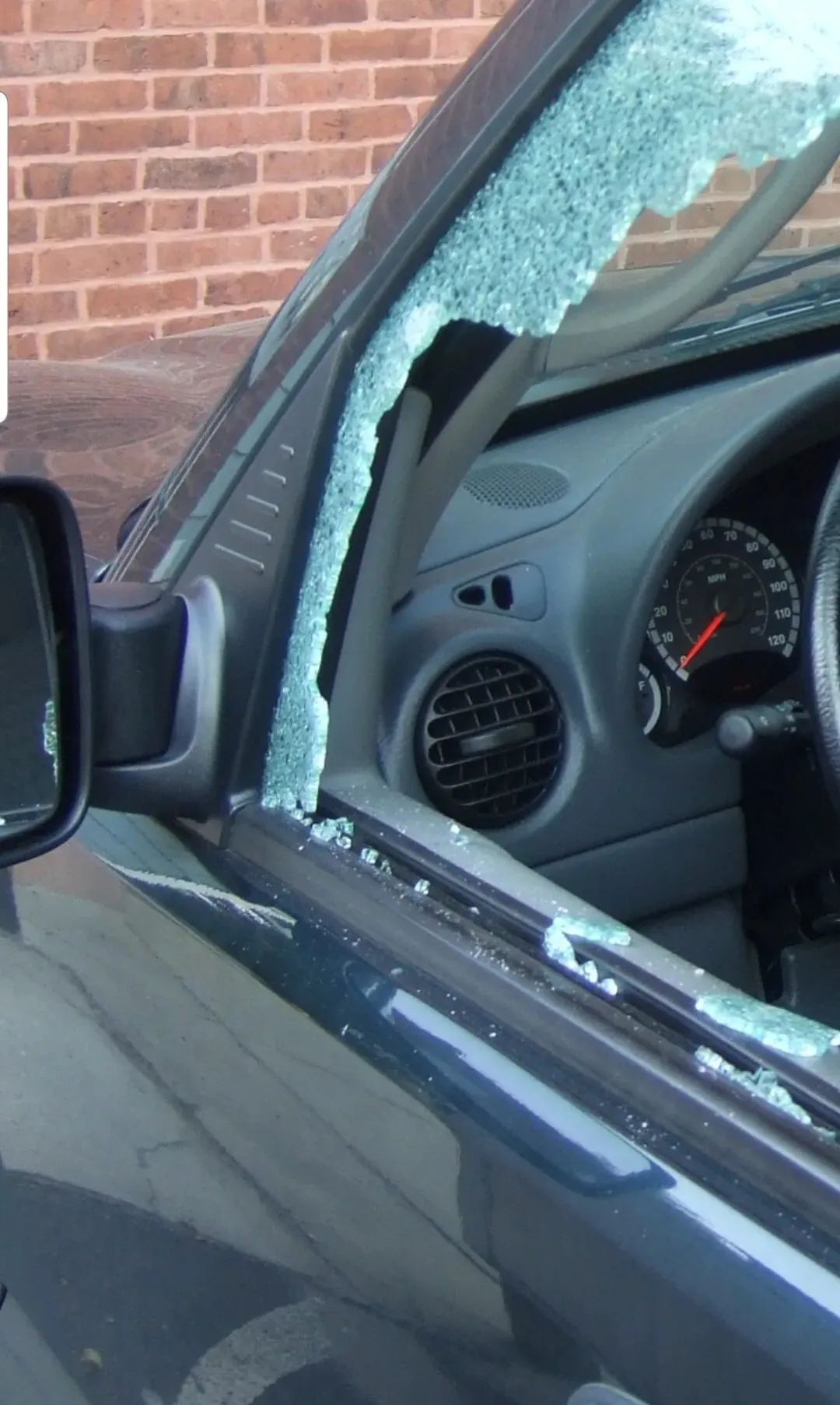 Car windshield shattered beside a brick wall, with broken glass on the window frame and dashboard visible inside