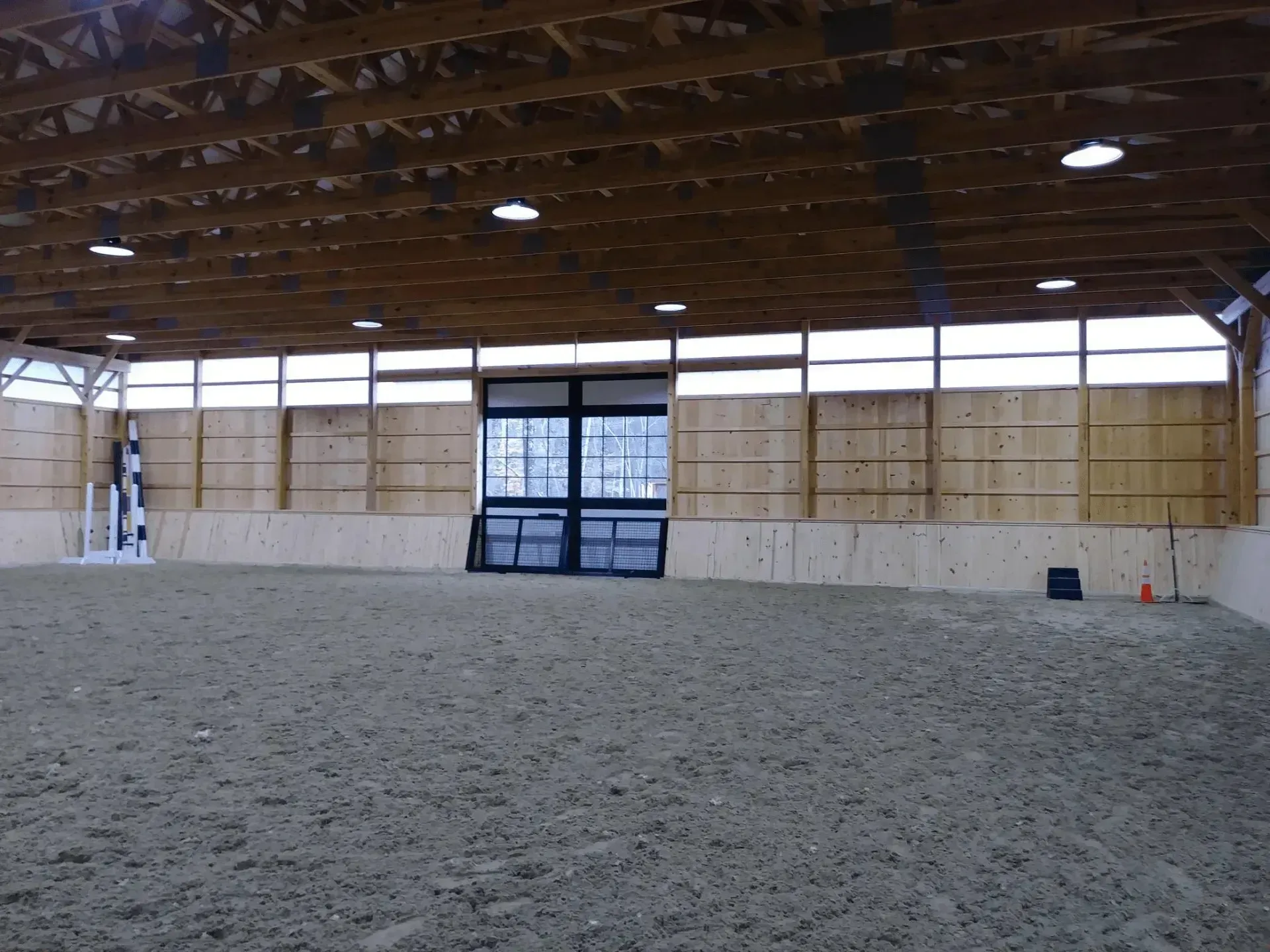 Indoor riding arena with sand footing, wooden walls, and large central doors.