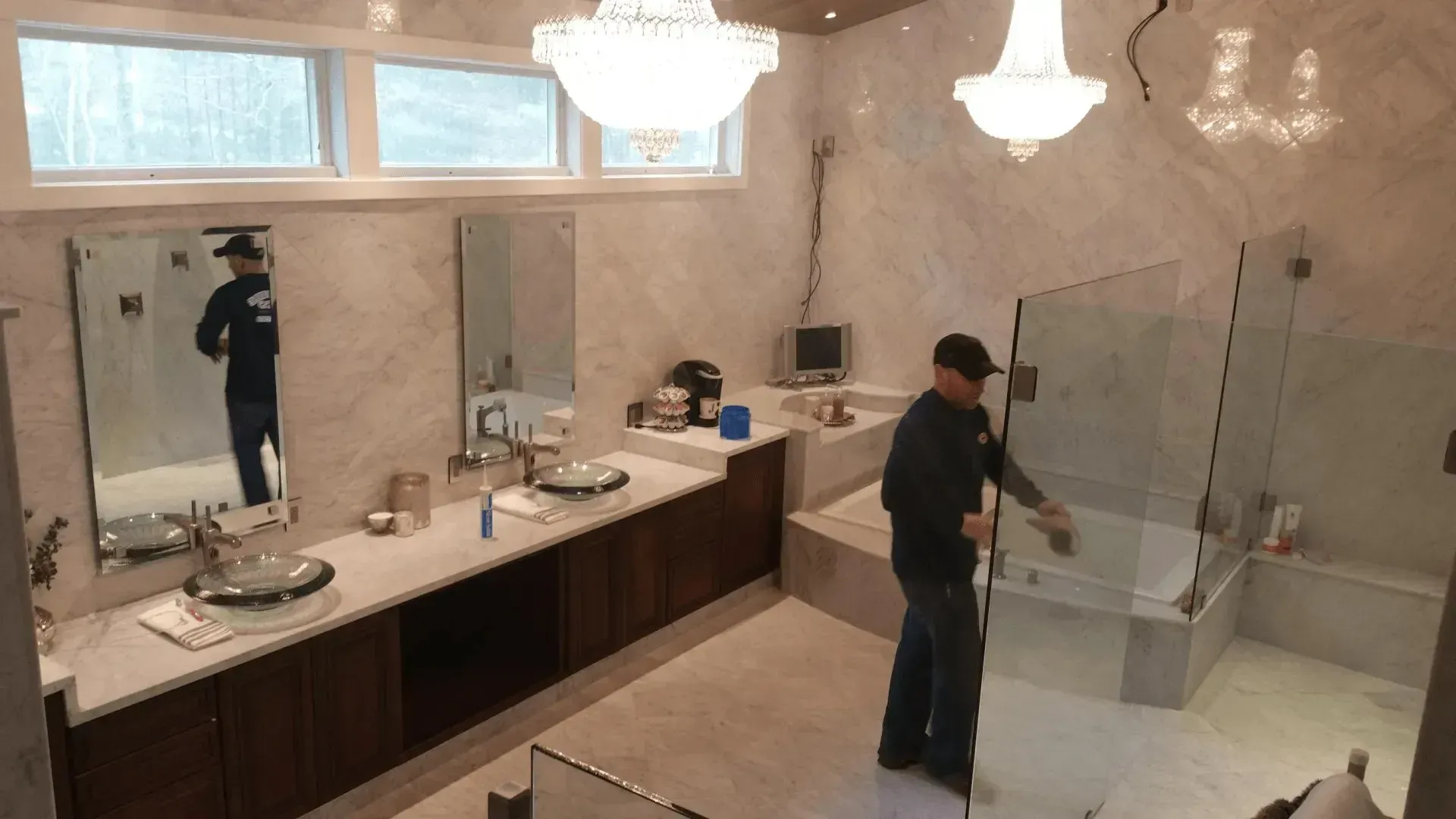 Spacious marble bathroom with dual sinks, large mirror, glass shower, and a person cleaning the floor.