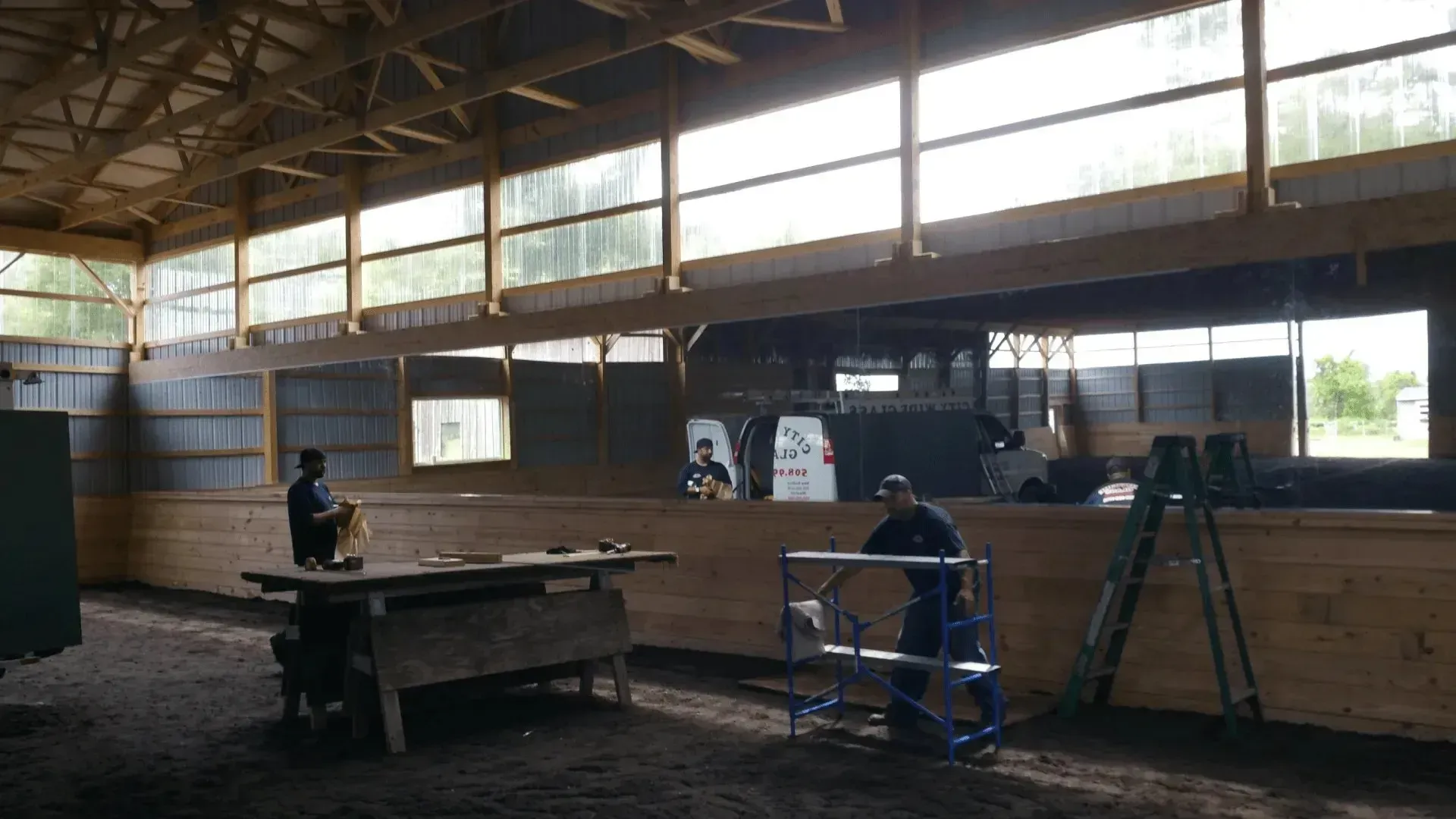 Spacious indoor warehouse with two people, worktables, and ladders under a high windowed roof.