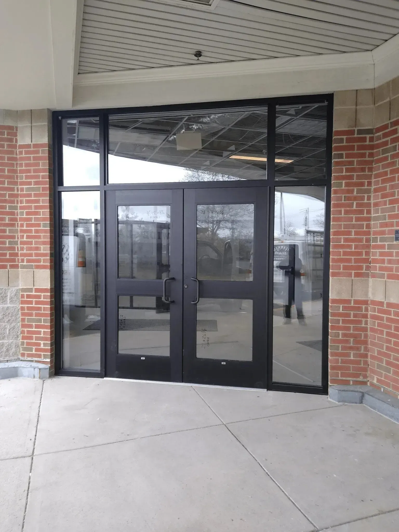 Black-framed glass double doors at a brick building entrance under a covered walkway.