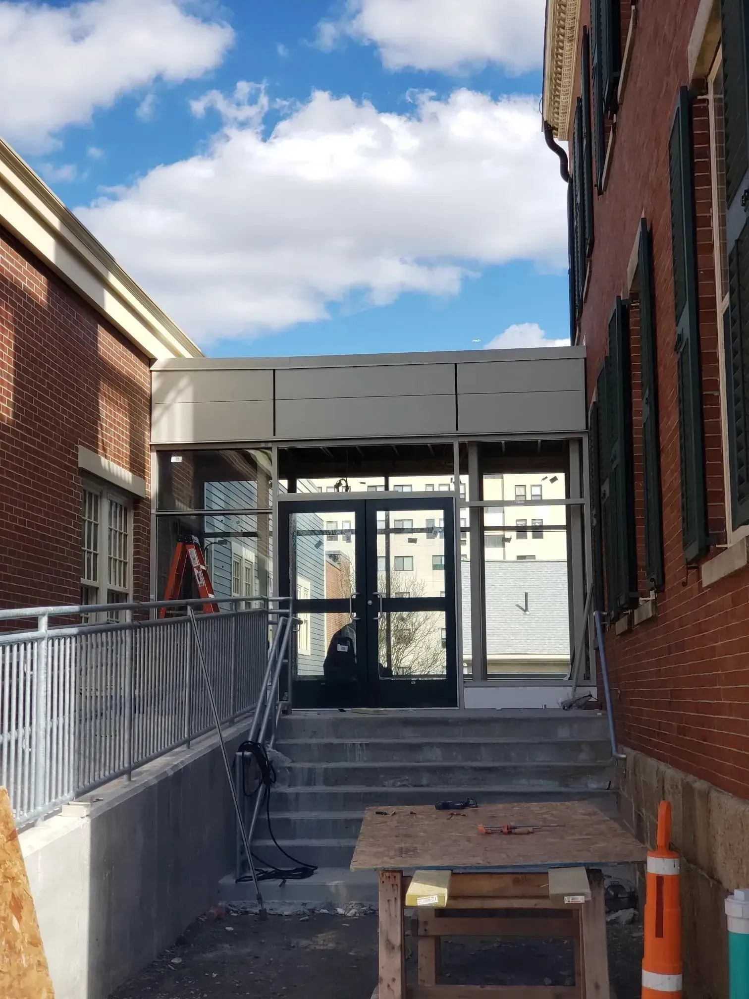 Narrow outdoor walkway between brick buildings leading to glass doors, with steps, ramp railing, and orange cone.