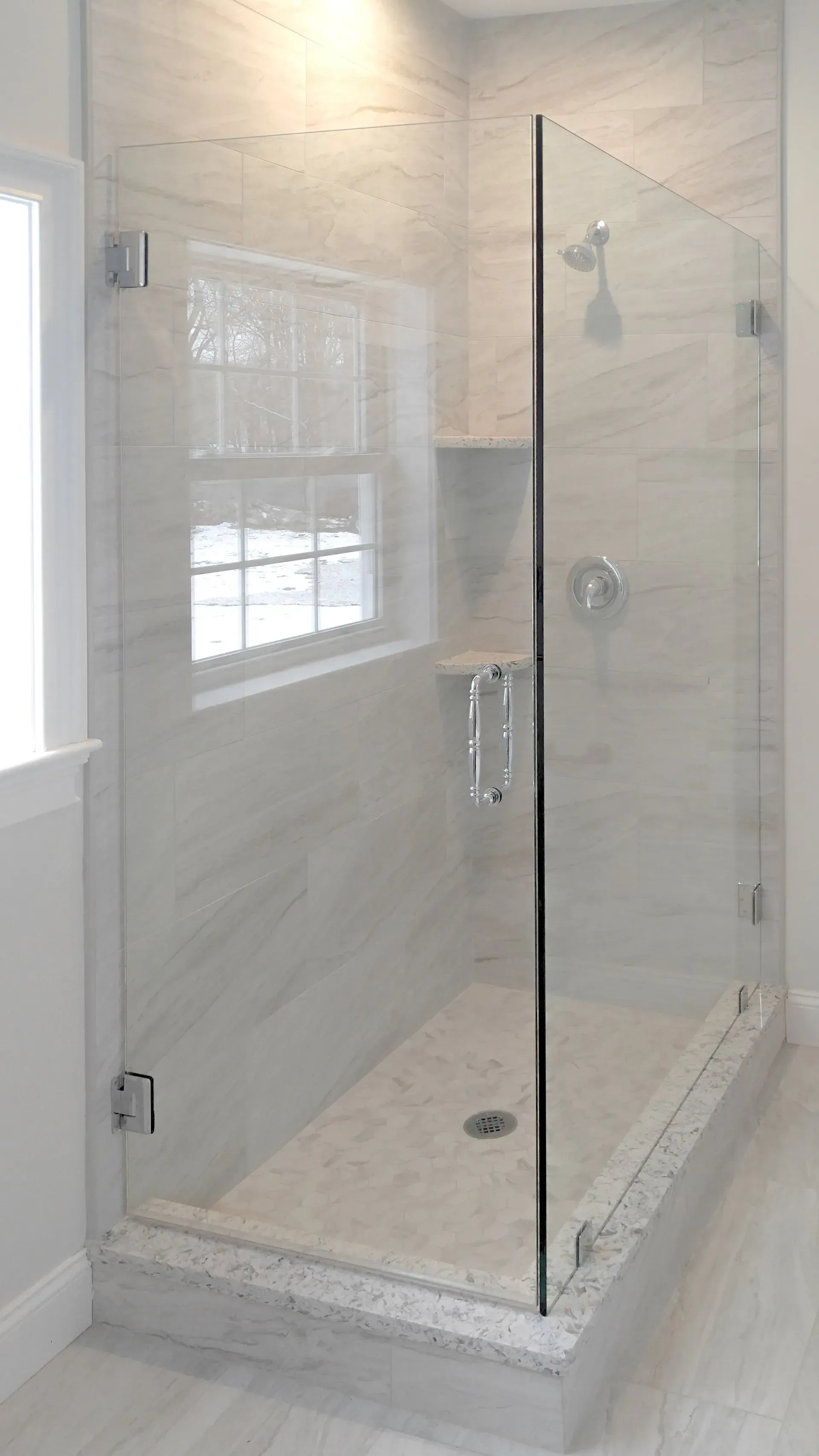 Glass walk-in shower with marble tile walls and a bench in a bright bathroom
