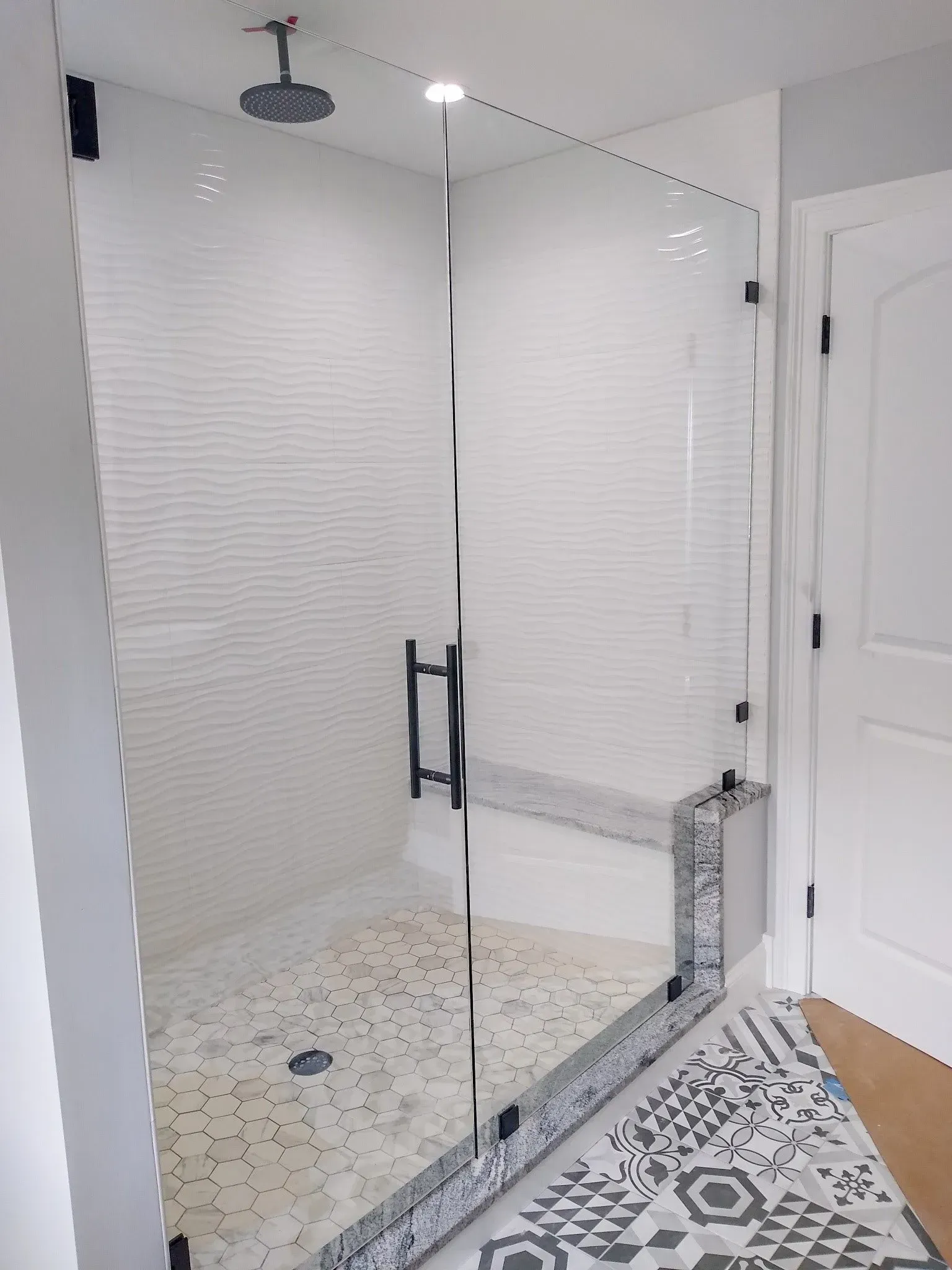 Glass shower enclosure with black handle in a white bathroom, beside patterned tile flooring and a white door