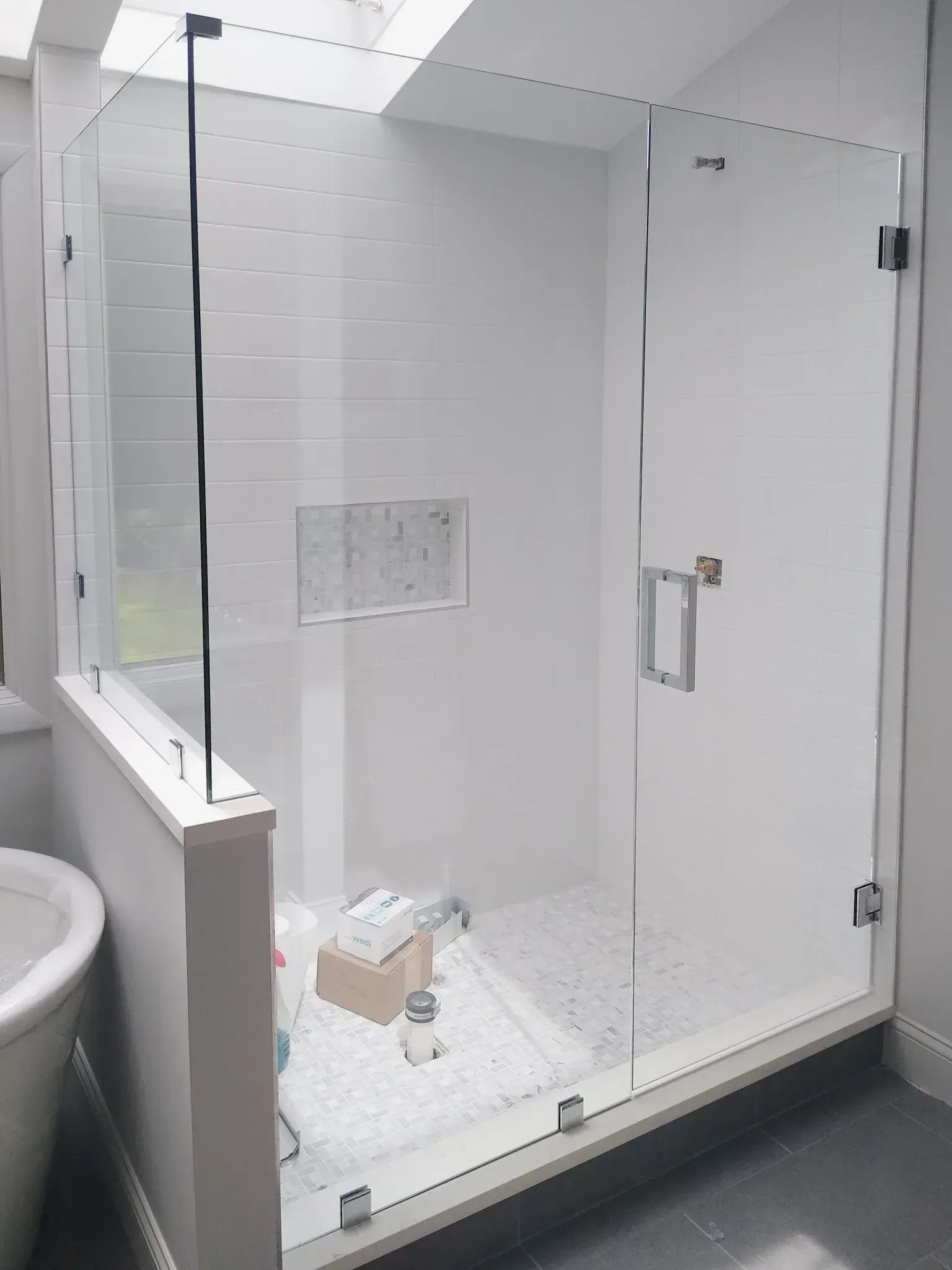 Small glass shower stall in a white tiled bathroom with a bench and shower fixtures, door partly open.