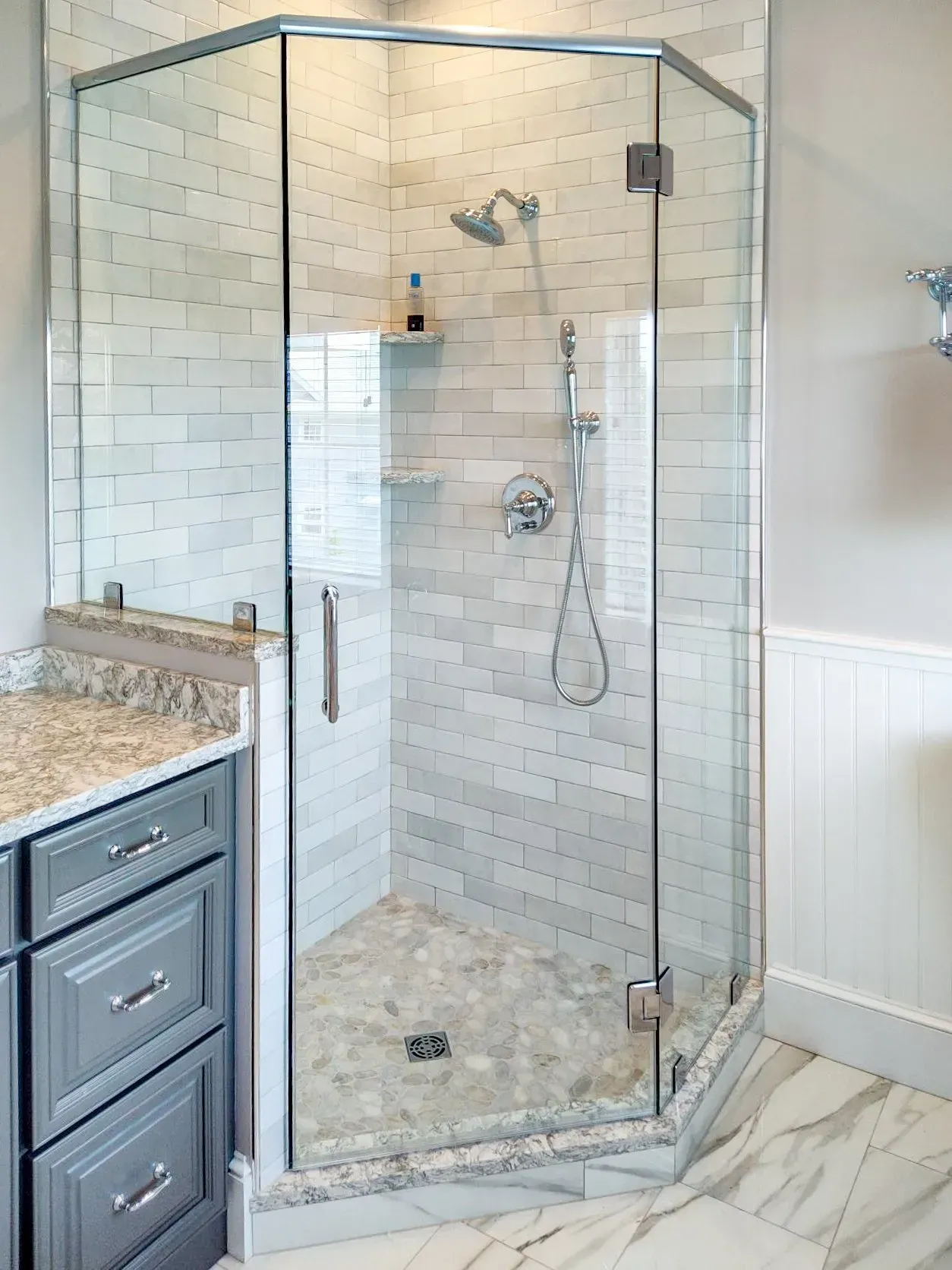 Corner glass shower with white tile, chrome fixtures, and a light granite bathroom vanity nearby