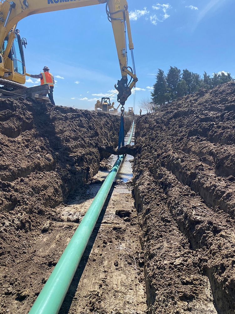 A yellow excavator is digging a trench with a green pipe