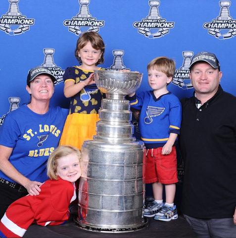 Family poses with the Stanley Cup, wearing St. Louis Blues apparel, in front of a celebratory backdrop.