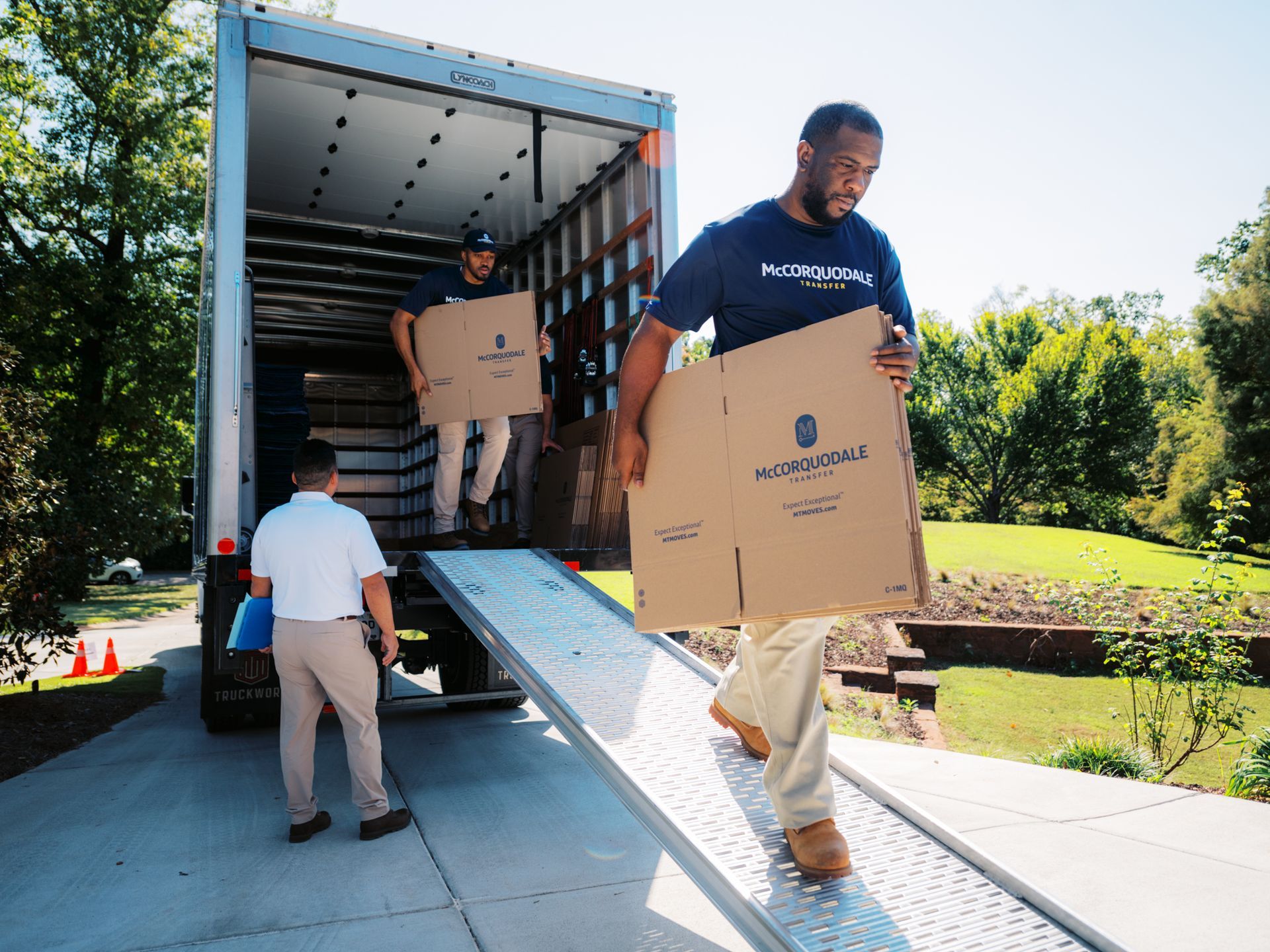 Movers unloading boxes from a truck onto a ramp, outdoors.