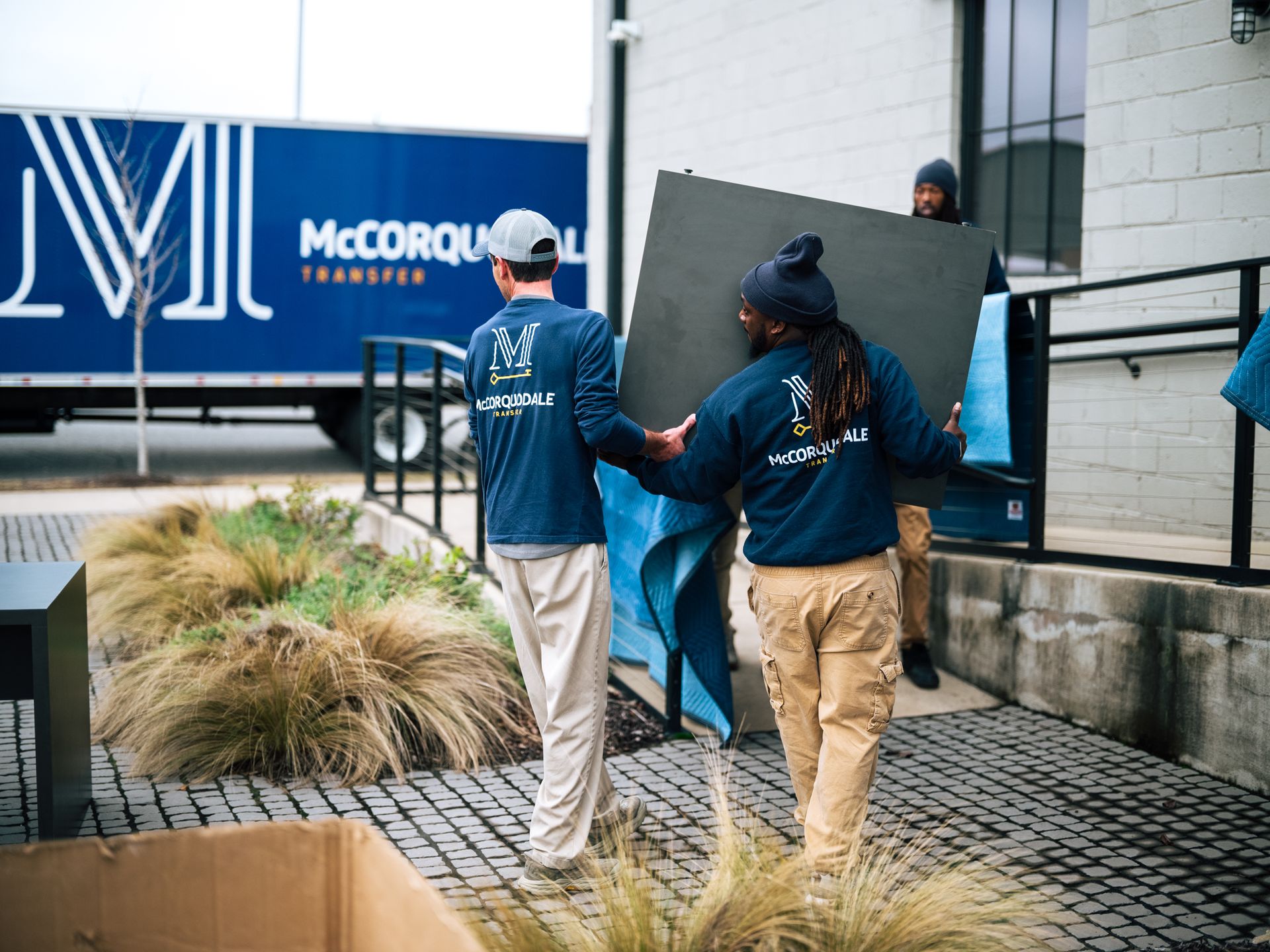 Movers carry a large, dark panel from a building to a moving truck. The truck is labeled