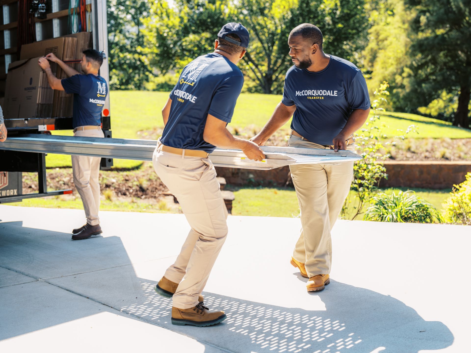 Movers carrying a large piece of furniture from a truck onto a driveway, other boxes being loaded. Sunny outdoors.