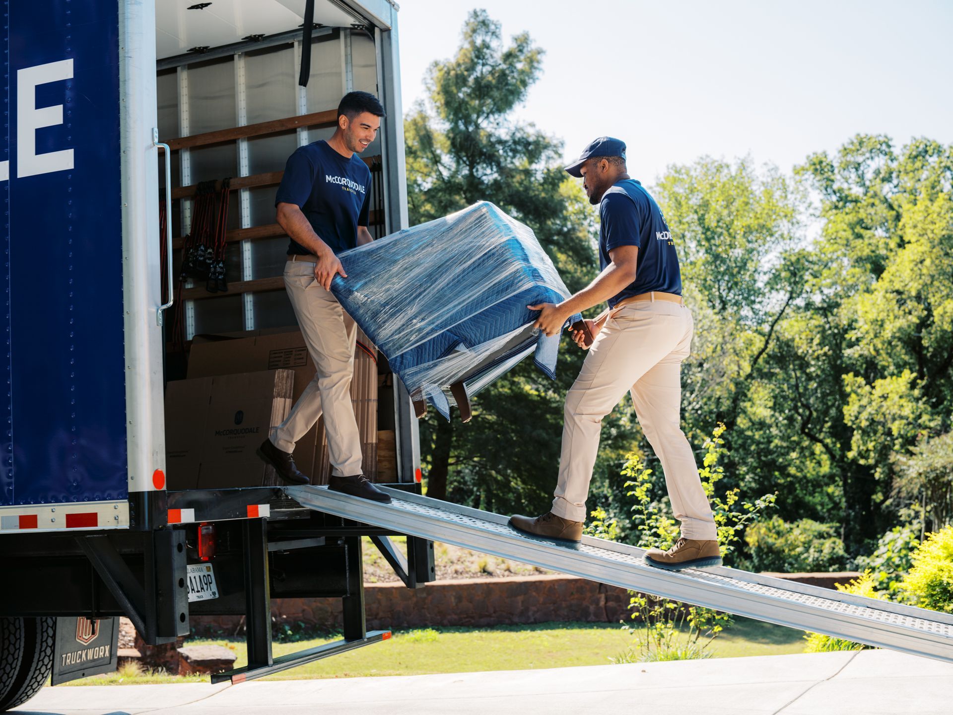 Two movers loading a blue-wrapped chair onto a truck via a ramp on a sunny day.