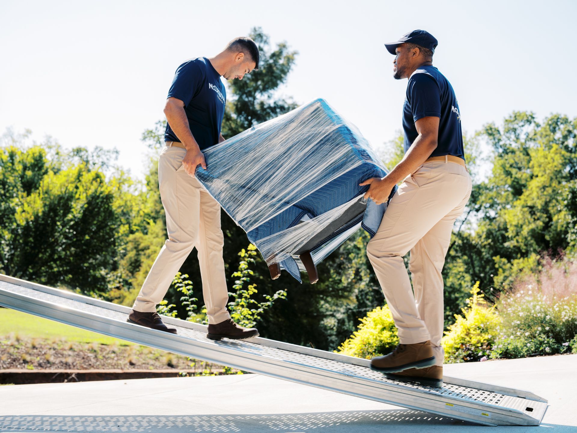 Two movers carrying wrapped furniture down a ramp outdoors.