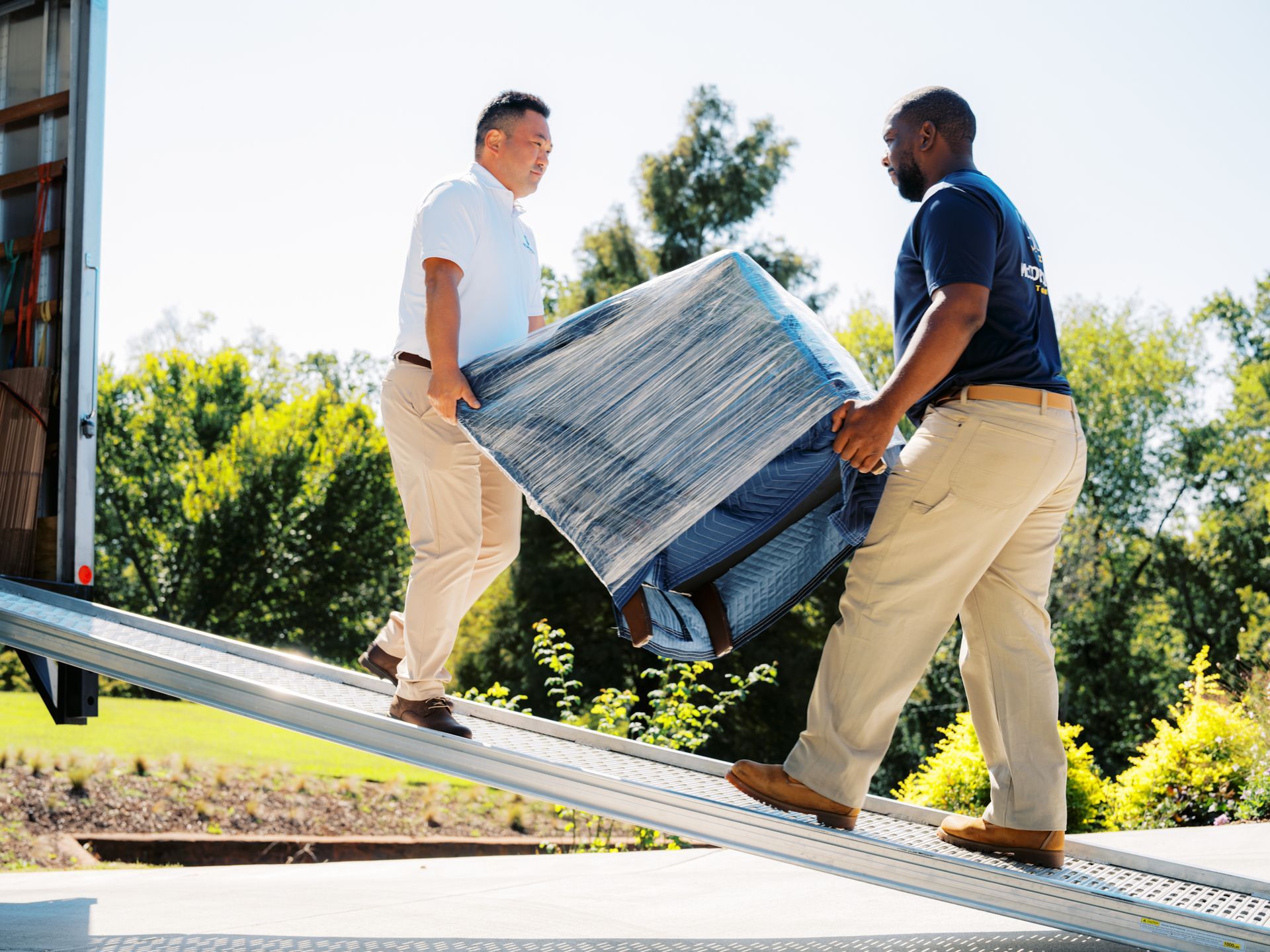 Two movers carrying wrapped furniture down a ramp into a truck on a sunny day.