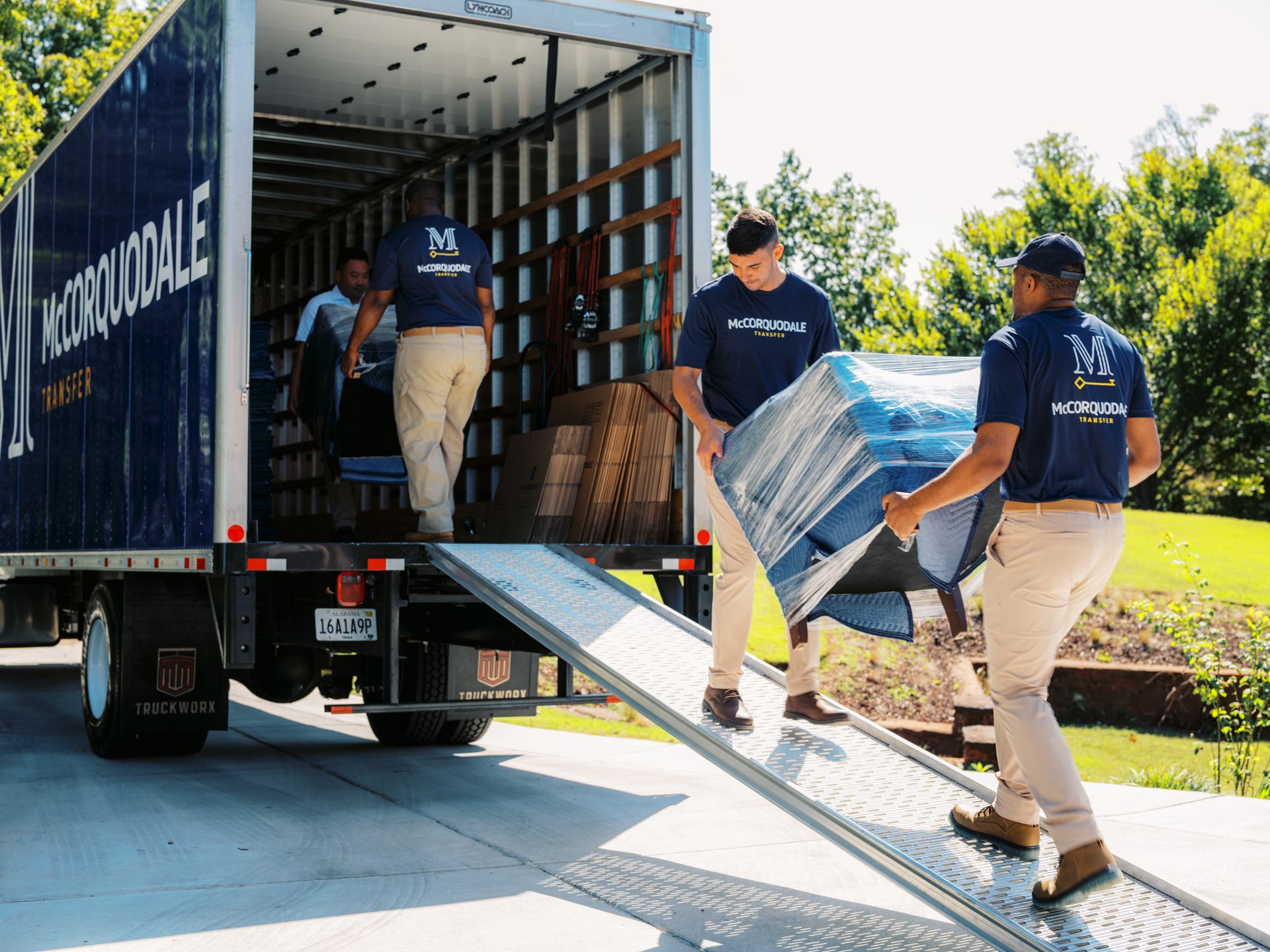 Movers loading a truck with a ramp, carrying a wrapped piece of furniture, outdoors.
