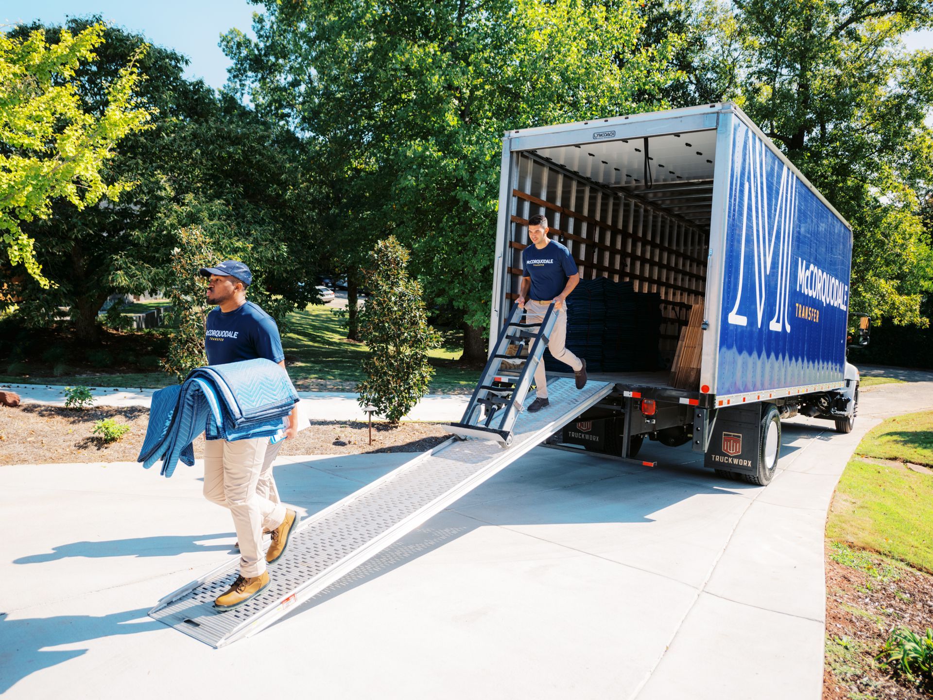 Two movers loading a moving truck on a driveway. One carries blue padding, the other uses ramp.