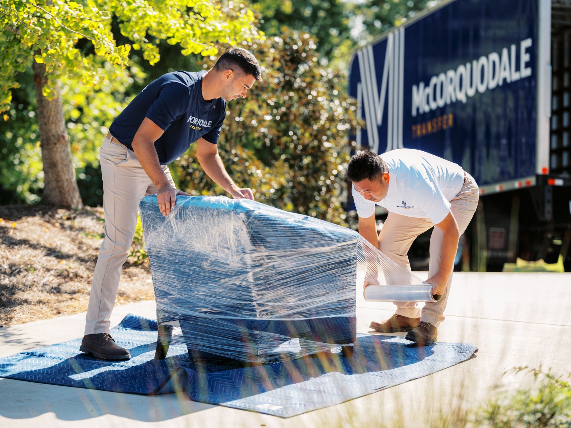 Two movers wrapping furniture in plastic next to a moving truck.