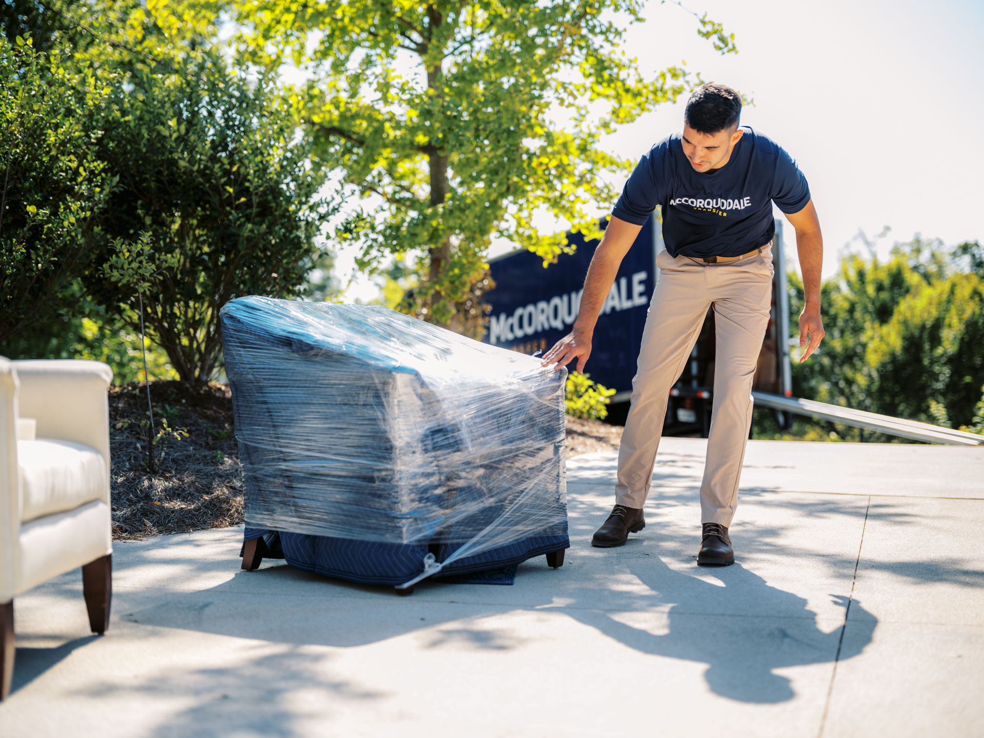 A person in a blue shirt and khaki pants rolls a wrapped chair onto a truck in a driveway.