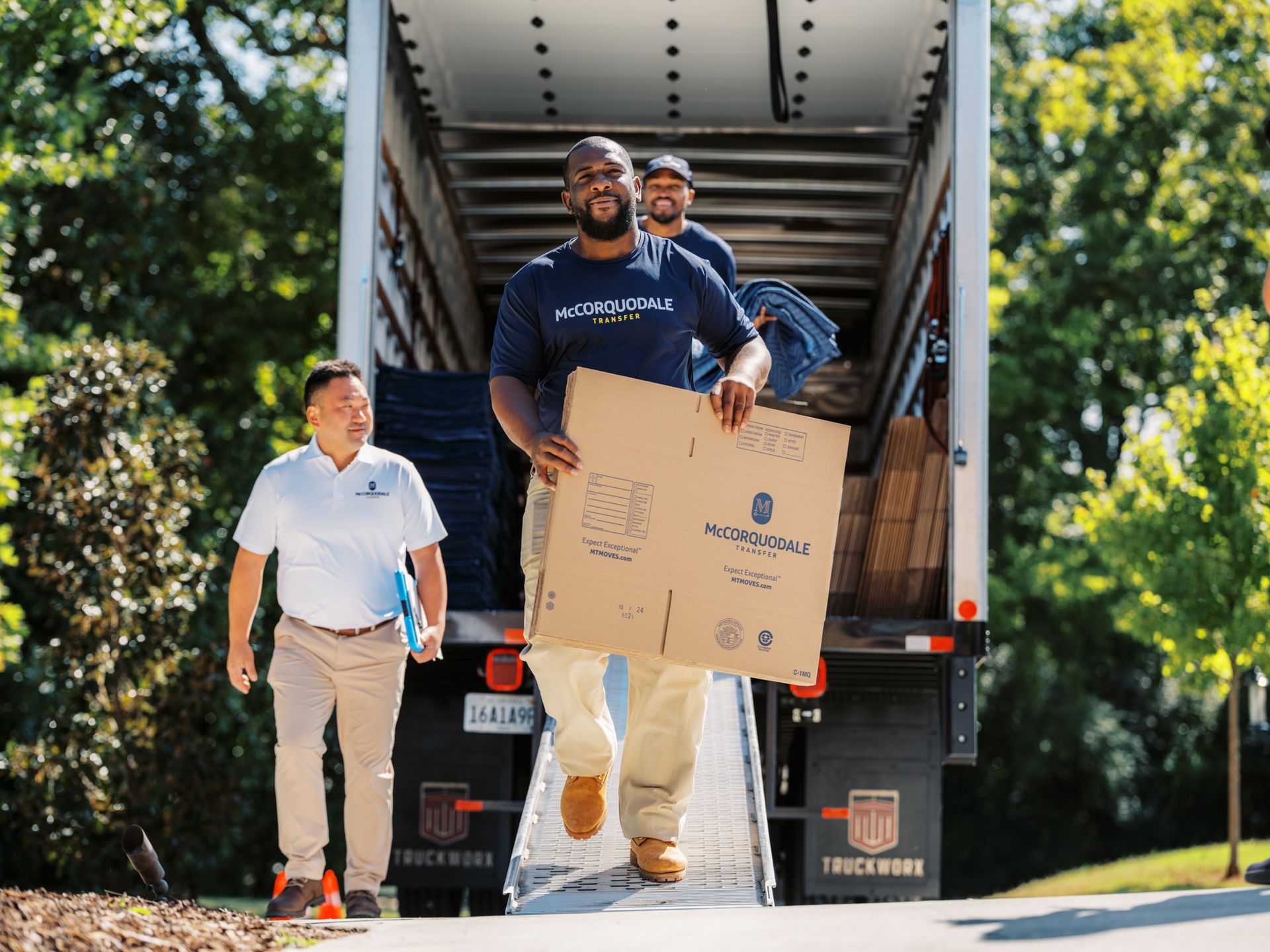 Movers unloading a cardboard box from a truck; one man smiling, other workers in the background.
